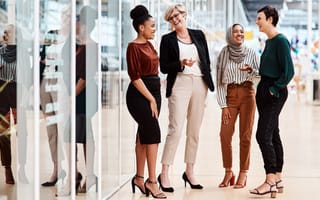 Four women colleagues laughing and chatting while wearing business attire in office hallway